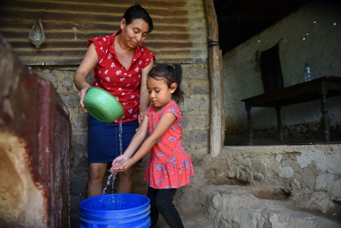 Eine Frau gießt aus einer Schüssel Wasser, daneben steht ein Kind und hält die Hände unter den Wasserstrahl, darunter eine blauer Tonne