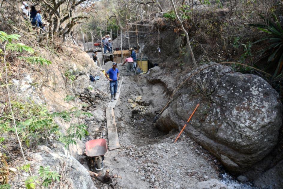 Ein ausgetrocknetes Flussbett, im Vordergrund ein großer Steinblock, Bretter, einige Arbeiter, Schubkarre