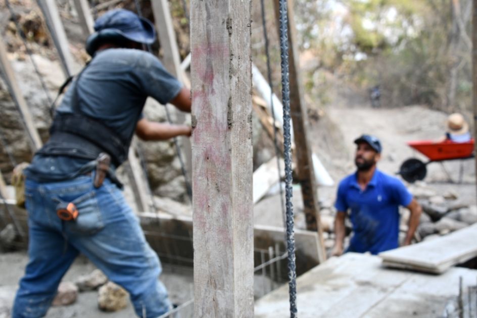 Zwei Männer mit großen Holzstämmen und Baumaterialien stehen in einem ausgetrocknetem Flussbett