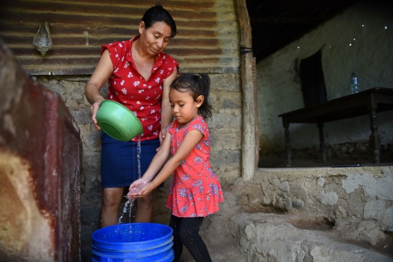 Auch beim Händewaschen muss Wasser sehr sparsam verwendet werden. Eine Frau gießt aus einer Schüssel Wasser, daneben steht ein Kind und hält die Hände unter den Wasserstrahl, darunter eine blauer Tonne