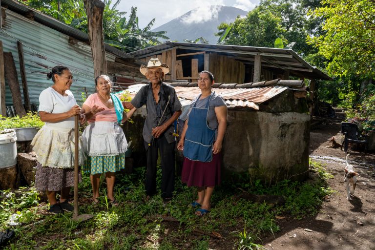 Wie viele Familien in Zentralamerika leben María Lavinia Quintanilla, Gloria Campos, Juan Antonio Moya und Dinora Gómez von dem was sie selbst anbauen. Mit Hilfe von OIKOS sind ihre Ernten reicher und vielfältiger geworden. Drei Frauen und ein Mann mit Hut stehen in El Salvador vor einem Haus. Eine Frau hat eine Hacke in der Hand.